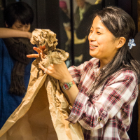 A woman with long hair, wearing a plaid shirt and a colorful wristwatch, joyfully crumples brown packing paper at the Papīra objektu festivāls. Behind her, another person with short hair in a blue shirt and dark scarf watches, covering their mouth with their arm. The background features a window reflecting figures in a room.