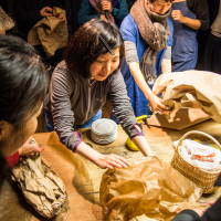A group of people are gathered around a wooden table, unwrapping and organizing ceramic plates from brown paper packages. One person in the center, wearing a gray jacket, is reaching for a stack of plates. Others are assisting and observing. The atmosphere resembles a festivāls of Papīra objekti, collaborative and focused.
