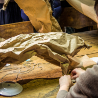 A person with short hair works on creating a sculpture using crumpled brown paper and a lamp base on a worn wooden table, as part of the Papīra objektu festivāls. Various craft materials and another partially seen individual are in the background, making for a lively and creative indoor setting.
