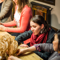Four people are seated around a wooden table engaged in crafting papīra objekti. One woman in the foreground wears a red scarf and closely examines the paper. Another, in a pink cardigan, focuses intently on her work. The room has a cozy, rustic feel with a window and radiator in the background.