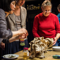 A group of four people stands around a wooden table. One person on the left is handling items wrapped in crumpled brūnais Papīra. Various objektu, including plates, a jar with yellow liquid, a canister, and dried leaves, are spread out. The mood appears to be focused and collaborative as if preparing for a festivāls.