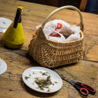 A wicker basket, partially filled with wrapped items, rests on a wooden table. Beside it, there is a yellow oil dispenser. In the foreground, a white plate holds herbs, and a pair of red-handled scissors lies nearby. A flyer for the Papīra objektu festivāls peeks out from behind the basket.