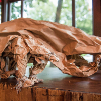 A sculpture of a bear made from crumpled brown paper is displayed on a wooden surface. The detailed paper folds create a lifelike representation of the bear, positioned against a background of large windows showcasing a green, leafy outdoor scene, evoking the essence of the Papīra objektu festivāls.