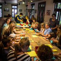 A group of adults and children gather around a long wooden table in a bright, well-lit room. Engaging in a craft activity for the festivāls, they are folding colorful papīra and creating art. The participants appear focused and engaged. The room has large windows, framed pictures, and light fixtures on the ceiling.