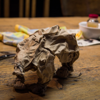 A crumpled paper sculpture of a bear stands on a wooden table. To the left, a pair of red-handled scissors lies open. In the background, various art supplies, including pencils, cloth, and a small jar, are scattered around, reminiscent of an art festivāls with wooden chairs visible further back.