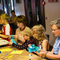 A group of children and adults are sitting around a wooden table at the festivāls, engaging in craft activities. They are folding colorful origami papers, transforming them into intricate papīra objekti. The table is scattered with paper pieces, tape, and craft tools. In the background, other people are observing. The scene is lively and creative.