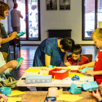 A group of children and adults are gathered around a table at the Papīra Objektu Festivāls, engaged in arts and crafts. Some are folding colored paper, while others assist the children. The room is well-lit with large windows in the background, allowing natural light to fill the space. Various supplies are scattered on the table.