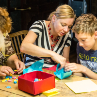 A woman in a striped shirt helps a boy in a blue t-shirt with an origami project during a papīra objektu festivāls. They are seated at a wooden table cluttered with colorful paper, and a red box is in front of them. Two other people, partially visible, are also engaged in folding paper on the left side of the image.