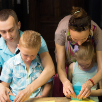A man and a woman assist two young children with an art project at a wooden table. They are folding and cutting colorful paper, perhaps preparing for the upcoming Papīra objektu festivāls. Scissors and other craft materials are scattered on the table. The adults are focused on helping the children, who appear engaged in the activity.