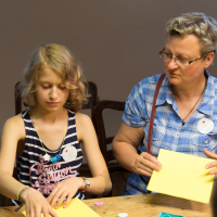 A young girl and an older woman are sitting at a wooden table engaged in a crafting activity, preparing for the upcoming festivāls. The girl, dressed in a striped tank top, is focused on her papīra objektu with small items, while the woman in blue plaid supervises, holding a yellow piece of paper.