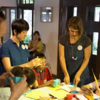 A group of children and adults is engaged in a crafts workshop inside a well-lit room with wooden furniture. Two adults assist the children, who are focused on colorful Papīra and craft materials on a large table. Various completed objektu projects are visible, and everyone looks absorbed in their activity.