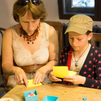 A woman and a child are engrossed in crafting papīra objektu at a wooden table. The woman, wearing sunglasses on her head, a light dress, and a beaded necklace, guides the child in a star-patterned cardigan who mirrors her actions. On the table are colorful papers, small boxes, and crafting tools.