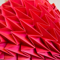 Close-up of an intricate red origami sculpture featured at the Papīra objektu festivāls, showcasing numerous sharp, angular folds and geometric patterns. The 3D paper artwork presents a complex, abstract form with layered triangular shapes creating a textured and vibrant surface.