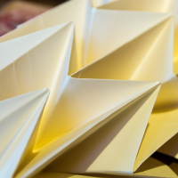 Close-up of intricately folded yellow Papīra origami creating a geometric, tessellated pattern. The folded shapes form sharp angles and interconnected points, casting soft shadows and creating a textured, three-dimensional appearance. The background is blurred and out of focus.
