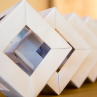 Close-up image of an abstract origami sculpture featuring a series of interconnected geometric shapes. Each shape consists of white paper folded into diamond-like frames, creating a repetitive, three-dimensional pattern. The background is blurred, focusing attention on the papīra objektu festivāls structure.