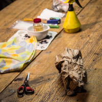 A wooden table with various items spread across: a pair of red and black scissors, a cloth with a fruit print, a crumpled brown Papīra bag, a yellow spray bottle, and a white bowl with small jars and plastic items next to a plastic bag. The background is softly lit, creating a warm ambience.