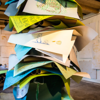 A large, colorful sculpture made of stacked, overlapping sheets of paper stands in an art gallery as part of the Papīra objektu festivāls. Each sheet features various doodles, drawings, and sketches in multiple colors. The artwork is displayed in a spacious room with wooden ceiling beams and unfinished walls adorned with additional drawings.