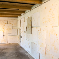 A gallery room with wooden ceiling beams displays numerous blank white canvases of various sizes, reminiscent of a Papīra objektu festival. The canvases are hung on white walls using clips and thin cords, creating a layered and overlapping effect. The concrete floor and lighting cast intriguing shadows on the canvases.