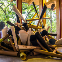 A large pile of cylindrical objects made of various materials and colors sits inside a room with floor-to-ceiling windows. Sunlight streams through the windows, illuminating the objects and casting shadows. Outside, green trees offer a vivid contrast to this creative indoor scene at the Papīra Objektu Festivāls.