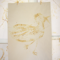 A drawing of a bird depicted using broad, flowing brushstrokes in sepia tones is displayed. The bird stands in profile on a large sheet of beige paper, which is secured to a line by two clothespins. Additional sheets with abstract designs hang around it like Papīra objektu festivāls, creating a gallery-like setting.
