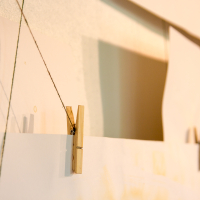 Close-up of two wooden clothespins clipped to a thin string, holding sheets of white papīra. The string is suspended horizontally, casting soft shadows on a beige background. The image is softly lit, highlighting the textures of the clothespins and the papīra.