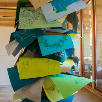 A towering art installation composed of large, colorful sheets of paper, layered and stacked haphazardly. Each sheet features intricate, hand-drawn sketches and diagrams. The structure sits in a wooden-floored gallery with large windows, allowing natural light to illuminate the display at the Papīra Objektu Festivāls.