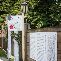 A pathway surrounded by green trees leads to a signboard displaying a map with several markings. To the right of the pathway, a wooden fence supports a white board featuring text in multiple languages about an upcoming festivāls. A vintage lamp post is positioned between the map and the text board.