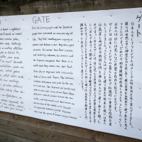 A bilingual exhibition panel featuring text in both European and East Asian languages is pinned to a wooden fence. The white panel with black text meticulously covers historical or informational content about "Gates" and Papīra objektu festivāls in the respective languages.