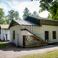 A small brick building with a metal roof and covered in white panels is situated in a park. The structure features several windows and an entrance. Surrounding the building are tall trees and grassy areas, hinting at the creativity of papīra māksla. A graffiti-covered wall is partially visible on one side. The sky is clear and sunny.