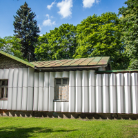 A small house with a corrugated metal exterior stands in a grassy area surrounded by tall, leafy trees. The roof appears weathered, showing signs of rust. Bright sunlight casts shadows on the house and the green grass. A path runs beside the house, reminiscent of routes taken during the Papīra objektu festivāls.
