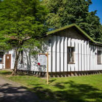 A white building with vertical paneling stands surrounded by greenery. A tree with dense foliage shades the left side, partially obscuring a door. A wooden signpost promoting the Papīra Objektu Festivāls is to the right of the door. The sky is clear and blue, casting sharp shadows on the grass and path in the foreground.