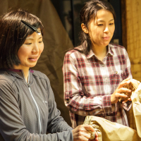 Two women are standing indoors, engaged in a task. The woman on the left is wearing a headlamp and a gray jacket, focused on an objektu in front of her. The woman on the right is in a red and gray plaid shirt, also looking down as she works, holding a brown papīra bag.