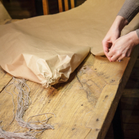 A pair of hands is seen wrapping a Papīra objektu in brown paper on a wooden table. Twine is spread out on the table, ready to be used for securing the package. In the background, there are wooden chairs, indicating a rustic setting. The scene suggests a process of packing or gift wrapping for an upcoming festivāls.
