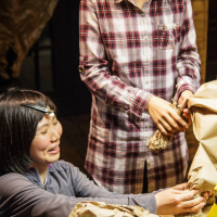 Two women are engaged in an arts and crafts activity, creating objektu sculptures with crumpled brown Papīra. One woman, standing, wears a plaid shirt and assists the other, who is seated in a gray top and wearing a headlamp. Several brown paper sculptures are on the table in front of them, ready for the upcoming festivāls.