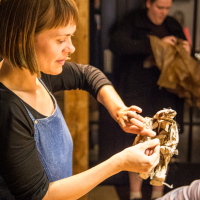 Woman in a denim apron smiling and shaping a piece of brown paper in her hands, with another person in the background also handling brown paper. They appear to be engaged in a Papīra craft activity, possibly preparing for an objektu festivāls, in a warmly lit indoor setting.