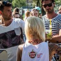 A crowd of people enjoying an outdoor event. At the foreground, a woman with platinum blonde hair and tattoos, wearing a white "Havana Club" top, is serving drinks. The man in front of her is speaking while holding a drink. Another man in sunglasses and a striped shirt stands nearby at Piens Fest 2014.
