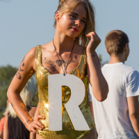 At the 2014 Piens Fest, a woman wearing a shiny gold sequin outfit holds a large white letter "R." With blonde hair styled in an updo and various tattoos on her arms and chest, she stands outdoors in sunny weather, while several people in casual clothing mingle in the background.