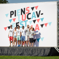 A group of seven people stands on a stage outdoors in front of a large backdrop that reads "Piens Lucav Festivāls 2014." The backdrop features colorful triangles and text in black, pink, and blue. The group is dressed casually in summer clothing, with most wearing sunglasses for the Piens Fest celebration.