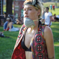 A person with light hair wears a feathered headpiece, a face mask hanging below their chin, a black bikini top, and an open, colorful sleeveless vest with intricate patterns. They are outdoors in a grassy area at Piens Fest 2014, with people sitting and relaxing in the background.