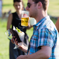 At Piens Fest 2014, a man balances a beer glass on his mouth while holding a wine glass and a plate of food in one hand at an outdoor event. Wearing a blue and white plaid shirt and sunglasses, he stands out as a woman can be seen in the blurred background, standing on the grassy area.