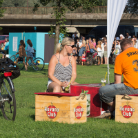 A woman and a man sit outdoors on Havana Club wooden crates, used as makeshift chairs, at Piens Fest 2014. The woman wears a striped tank top and sunglasses, holding a drink. The man, in an orange shirt with "BRACHA 33" on the back, faces her. Bicycles and people are visible in the background with a bridge overhead.