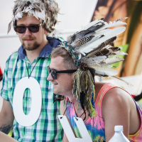 Two individuals wearing feathered hats and large white letters "O" and "H" around their necks sit at a table during Piens Fest 2014. The person on the left wears a green plaid shirt and sunglasses. The person on the right, in a tie-dye shirt and sunglasses, holds a water bottle. They appear to be part of an event.