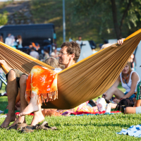 A man is lying in a brown hammock, reading a book at Piens Fest 2014, with an orange and yellow patterned blanket over his legs. People are sitting on the grass around him, some on blankets. Bicycles and a black vehicle are in the background. It's a sunny day with green grass and trees visible.