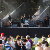 At the 2014 Piens Fest, an outdoor concert where a band performs on a stage, the female singer in a black dress captivates the audience. With a guitarist, drummer, and keyboardist by her side, the lively atmosphere is enhanced by bright sunlight as attendees in casual summer attire enjoy the show seated on the grass.