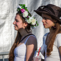 Two young women with long brown hair stand side by side at the Piens Fest 2014. One wears a white dress and a floral crown with white and pink flowers. The other wears a brown hat adorned with red cherries and a white dress, holding a drink with a red straw. They are outdoors in front of a white background.