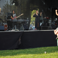 At Piens Fest 2014, a band performed on an outdoor stage with musicians playing instruments. In the audience, a person sat on another's shoulders, both raising their arms in enthusiasm. The scene was set on a sunny day with green grass in the area.