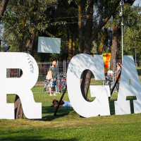 A large, white, 3D "RĪGA" sign is set up in a green park with scattered trees. People are sitting and relaxing on the grass near the sign. In the background, bicycles are parked against a metallic fence, and others walk or stand, enjoying the sunny day of Piens Fest 2014.