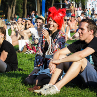 At PiensFest 2014, a group of people is sitting on the grass at an outdoor event on a sunny day. They appear to be enjoying the fest, with some smiling and laughing. One person in the center stands out with bright red hair and colorful clothing. Trees and other attendees are visible in the background.
