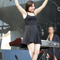 At Piens Fest 2014, a woman in a black dress energetically holds a microphone in one hand and raises her other arm while standing on an outdoor stage. Another woman smiling and dressed in black stands near a keyboard in the background. Stage equipment and a performer playing guitar are partially visible.