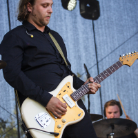 A musician with shoulder-length hair plays an electric guitar on stage at Piens Fest 2014. He wears a black shirt and has a strap over his shoulder. In the background, another musician is visible playing drums. Stage lights and draped netting create ambiance in the setting.
